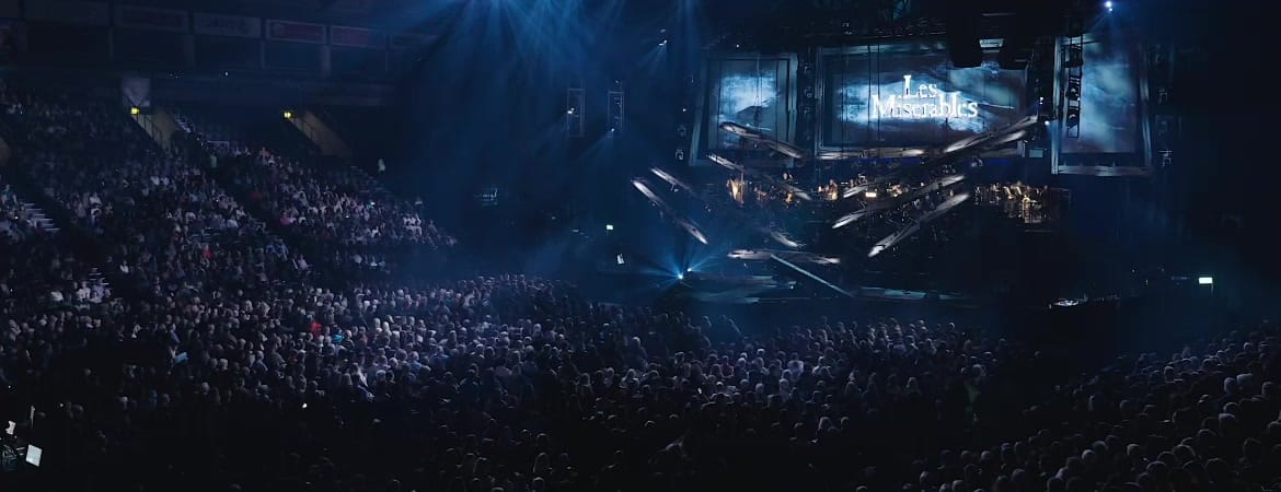 A wide arena view of Les Misérables in concert: the full stage rig over a capacity crowd